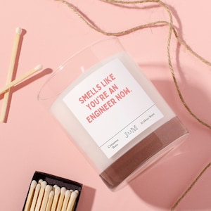 May include: A clear glass candle jar with a white label that reads "Smells like you're an engineer now." The candle is a cinnamon bun scent and has a 30-hour burn time. The candle is on a pink surface with a brown twine string and matches.