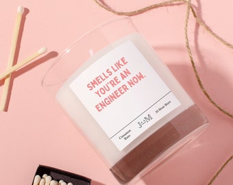 May include: A clear glass candle with white wax and a label that reads "SMELLS LIKE YOU'RE AN ENGINEER NOW." The label also includes the text "JAAM" and "30 Hour Burn" and "Cinnamon Buns." Two wooden matches and a matchbox are visible.