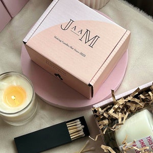Puede incluir: Una caja rosa con el texto "JAM Making Candles Hot Since 2020" en la parte delantera. La caja est&aacute; sobre una superficie rosa. Hay una vela blanca en un tarro de vidrio y una caja de cerillas en la superficie.