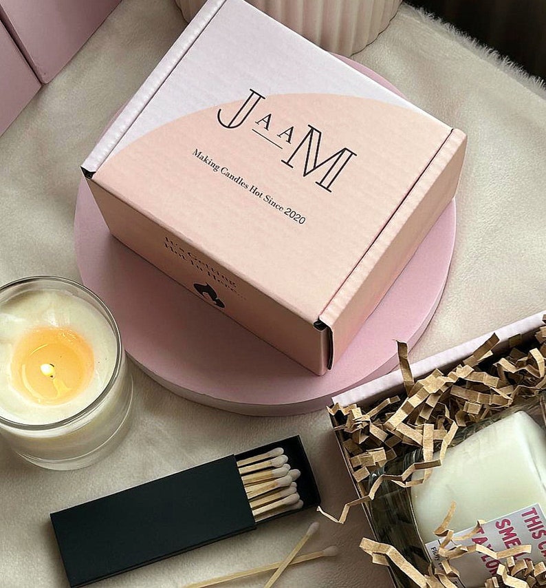 Puede incluir: Una caja rosa con el texto "JAM Making Candles Hot Since 2020" en la parte delantera. La caja est&aacute; sobre una superficie rosa. Hay una vela blanca en un tarro de vidrio y una caja de cerillas en la superficie.