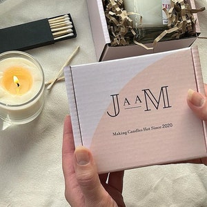 以下が含まれることがあります： ピンクと白の箱に「J A A M Making Candles Hot Since 2020」の文字が書かれています。箱は人の手で持たれています。