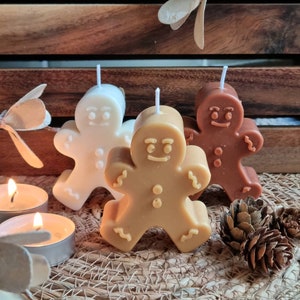 May include: Three gingerbread man shaped candles, one white, one brown, and one tan. The candles are on a woven surface with pine cones.