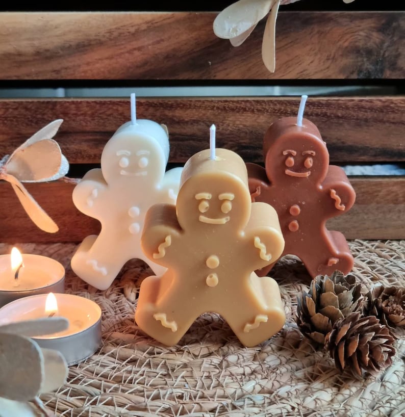 May include: Three gingerbread man shaped candles, one white, one brown, and one tan. The candles are on a woven surface with pine cones.