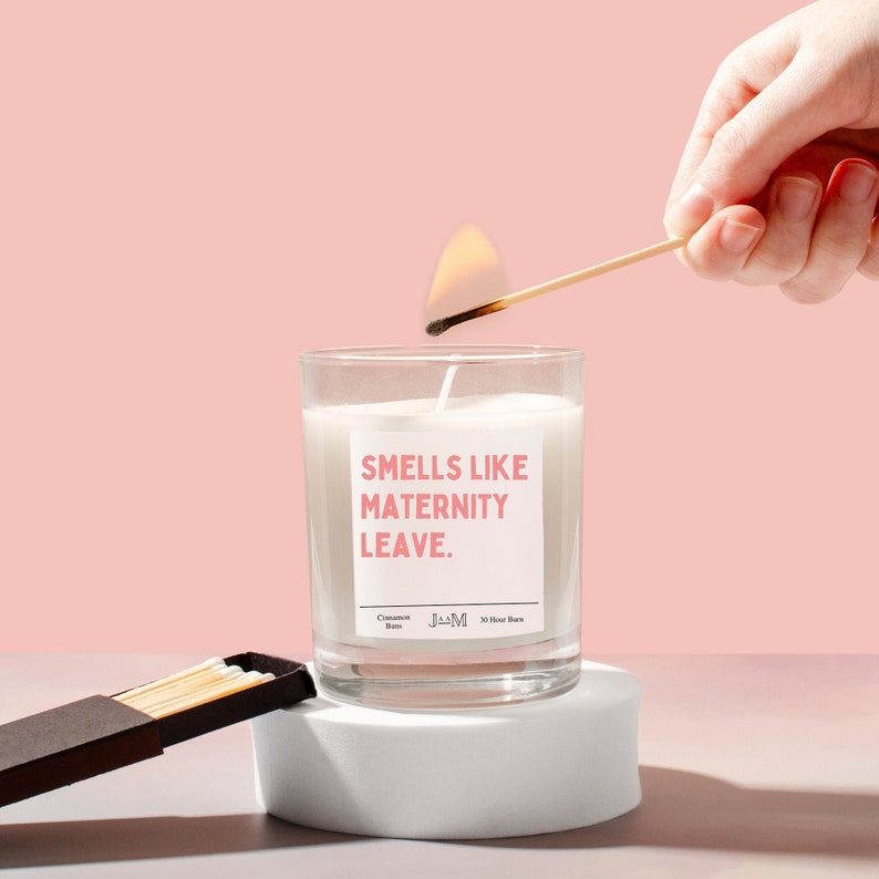 May include: A lit candle in a clear glass jar with the text "Smells Like Maternity Leave." A match is being used to light the candle. The candle sits on a white pedestal with a pink background. A box of matches is next to the pedestal.