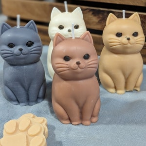 May include: Four cat-shaped candles in different colors: gray, white, brown, and tan. The candles are sitting on a blue surface.