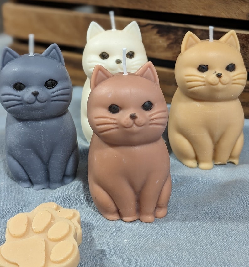 May include: Four cat-shaped candles in different colors: gray, white, brown, and tan. The candles are sitting on a blue surface.