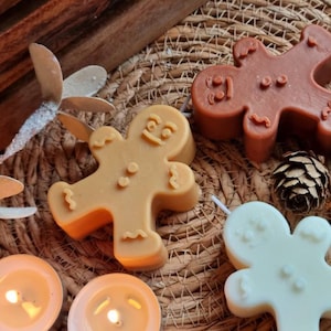 May include: Three gingerbread man shaped candles, one brown, one beige, and one white, on a woven placemat with two lit tea lights.