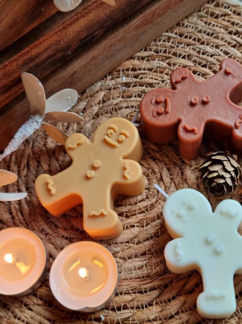 May include: Three gingerbread man shaped candles, one brown, one beige, and one white, on a woven placemat with two lit tea lights.