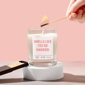 May include: A glass candle with the text "Smells Like You're Married" on a pink label. A match is being used to light the candle. The candle is on a white pedestal against a pink backdrop. The label reads "Cinnamon Buns" and "30 Hour Burn."