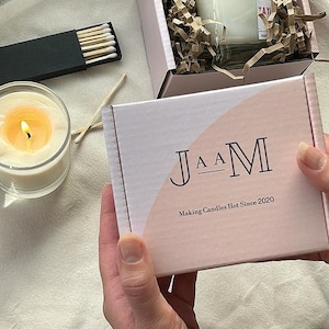 Puede incluir: Una caja blanca con un dise&ntilde;o rosa y blanco. La caja tiene el texto "J A A M" y "Making Candles Hot Since 2020" impreso en ella.