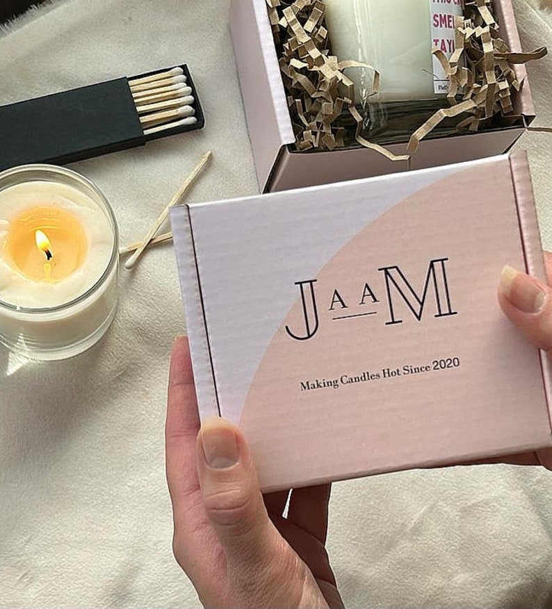 Puede incluir: Una caja blanca con un dise&ntilde;o rosa y blanco. La caja tiene el texto "J A A M" y "Making Candles Hot Since 2020" impreso en ella.