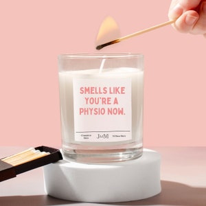 May include: A white candle in a glass jar with a pink label that reads "Smells like you're a physio now." The candle is lit with a match. The candle is on a white surface.