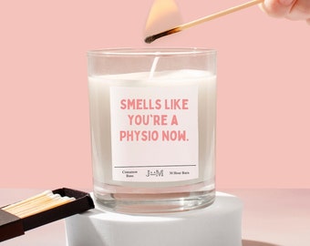 May include: A clear glass candle with white wax and a label that reads "SMELLS LIKE YOU'RE A PHYSIO NOW." The label also includes the text "Cinnamon Buns," "JAAM," and "30 Hour Burn." A lit match is held above the candle wick.