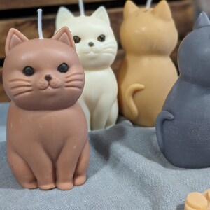 May include: Four hand-poured cat-shaped candles in brown, white, orange, and gray. The candles are sitting on a gray fabric surface.