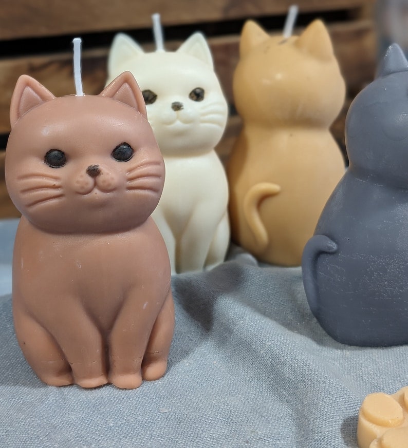 May include: Four hand-poured cat-shaped candles in brown, white, orange, and gray. The candles are sitting on a gray fabric surface.