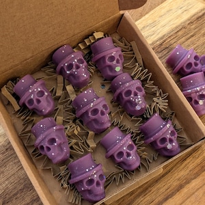 May include: A collection of purple skull-shaped wax melts, each adorned with a top hat and silver glitter. The melts are presented in a cardboard box with brown paper, ideal for Halloween or gothic-themed decorations.