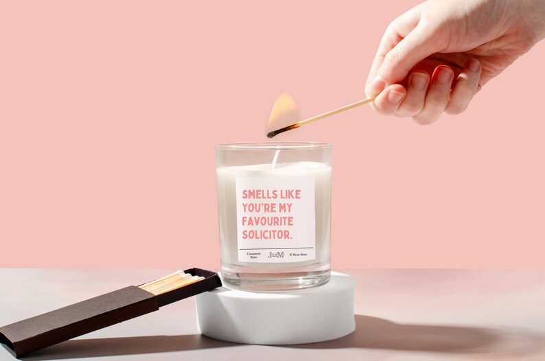 Puede incluir: Una vela blanca en un tarro de vidrio transparente con una etiqueta rosa que dice "Smells like you're my favourite solicitor." Una mano est&aacute; encendiendo la vela con una cerilla. Una caja de cerillas est&aacute; sobre una superficie blanca.