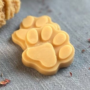 May include: Two beige paw-shaped soap bars with a raised paw print design. The soap bars are on a light gray textured surface.