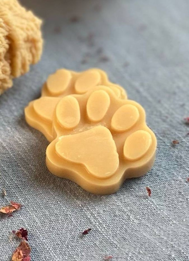 May include: Two beige paw-shaped soap bars with a raised paw print design. The soap bars are on a light gray textured surface.