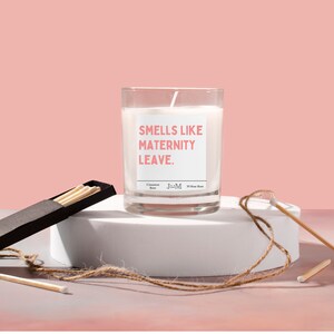 May include: A clear glass candle with the text "Smells Like Maternity Leave" in pink. The candle sits on a white pedestal with a box of matches and a piece of twine. The background is a solid pink.