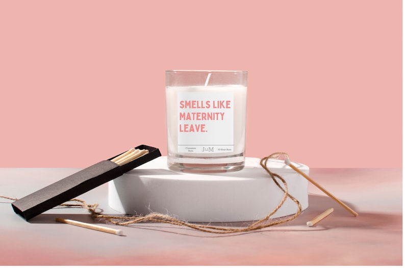 May include: A clear glass candle with the text "Smells Like Maternity Leave" in pink. The candle sits on a white pedestal with a box of matches and a piece of twine. The background is a solid pink.