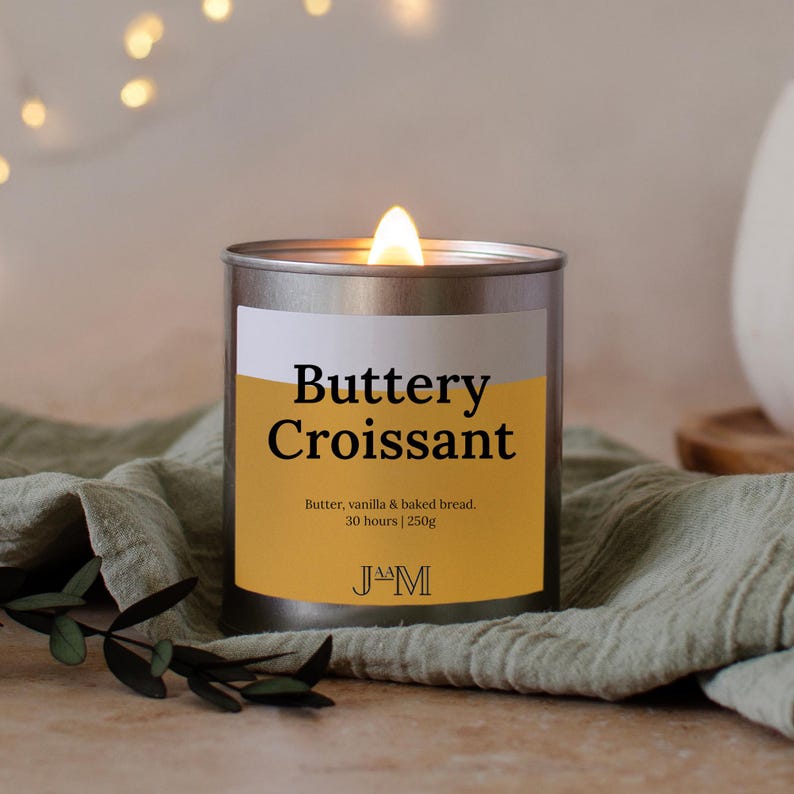 K&ouml;nnte beinhalten: Eine brennende Kerze in einer silbernen Dose. Das Etikett lautet "Buttery Croissant" und listet Butter, Vanille und gebackenes Brot auf. Sie hat eine Brenndauer von 30 Stunden und wiegt 250g. Die Kerze steht auf einem gr&uuml;nen Tuch.
