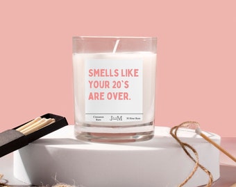 May include: A clear glass candle with white wax and a label that reads: SMELLS LIKE YOUR 20'S ARE OVER. Cinnamon Buns, JAAM, 30 Hour Burn. The candle is accompanied by a black matchbox containing wooden matches and loose twine.