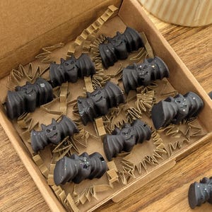 May include: A cardboard box filled with dark gray bat-shaped wax melts. The bats have detailed wings and smiling faces. The box is lined with brown crinkle paper, and the background is a wooden surface.