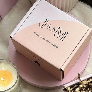 May include: A pink box with the text "JAAM Making Candles Hot Since 2020" on the front. The box is sitting on a pink surface.
