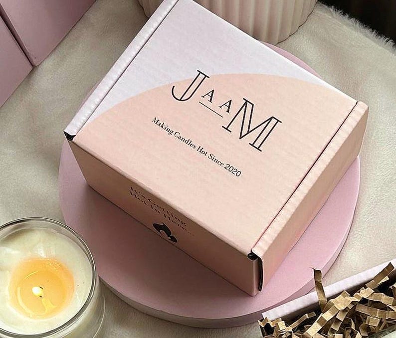 May include: A pink box with the text "JAAM Making Candles Hot Since 2020" on the front. The box is sitting on a pink surface.