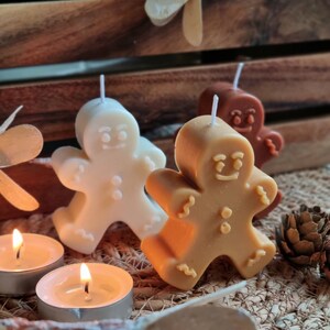 May include: Three gingerbread man-shaped candles, one white, one brown, and one tan, with wicks sticking out of the top. The candles are sitting on a woven brown surface with two lit tea lights.