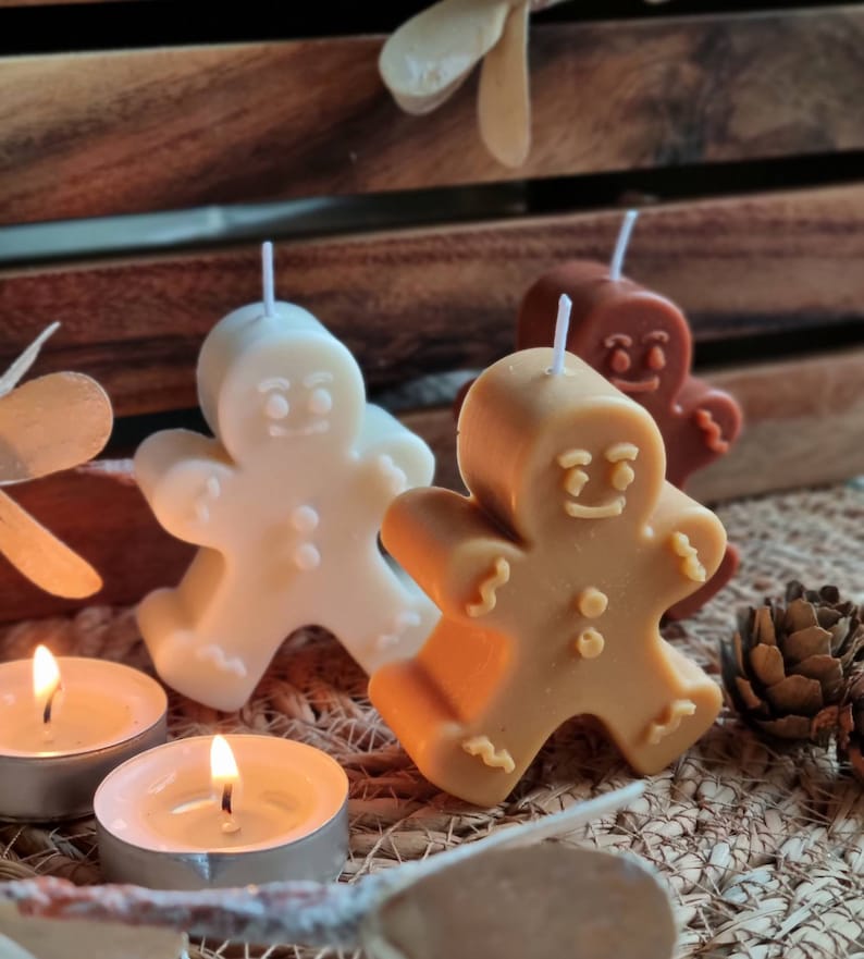 May include: Three gingerbread man-shaped candles, one white, one brown, and one tan, with wicks sticking out of the top. The candles are sitting on a woven brown surface with two lit tea lights.