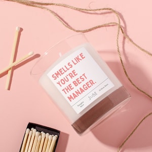 Puede incluir: Un tarro de cristal transparente con una vela y una etiqueta blanca que dice "Smells like you're the best manager." La etiqueta está en texto rosa con un contorno negro. La vela está sobre una superficie rosa con un cordel de yute marrón y dos cajas de cerillas.