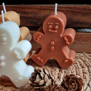 May include: Three gingerbread man shaped candles, one is brown, one is white, and one is tan. The candles are sitting on a woven basket surface with pine cones.
