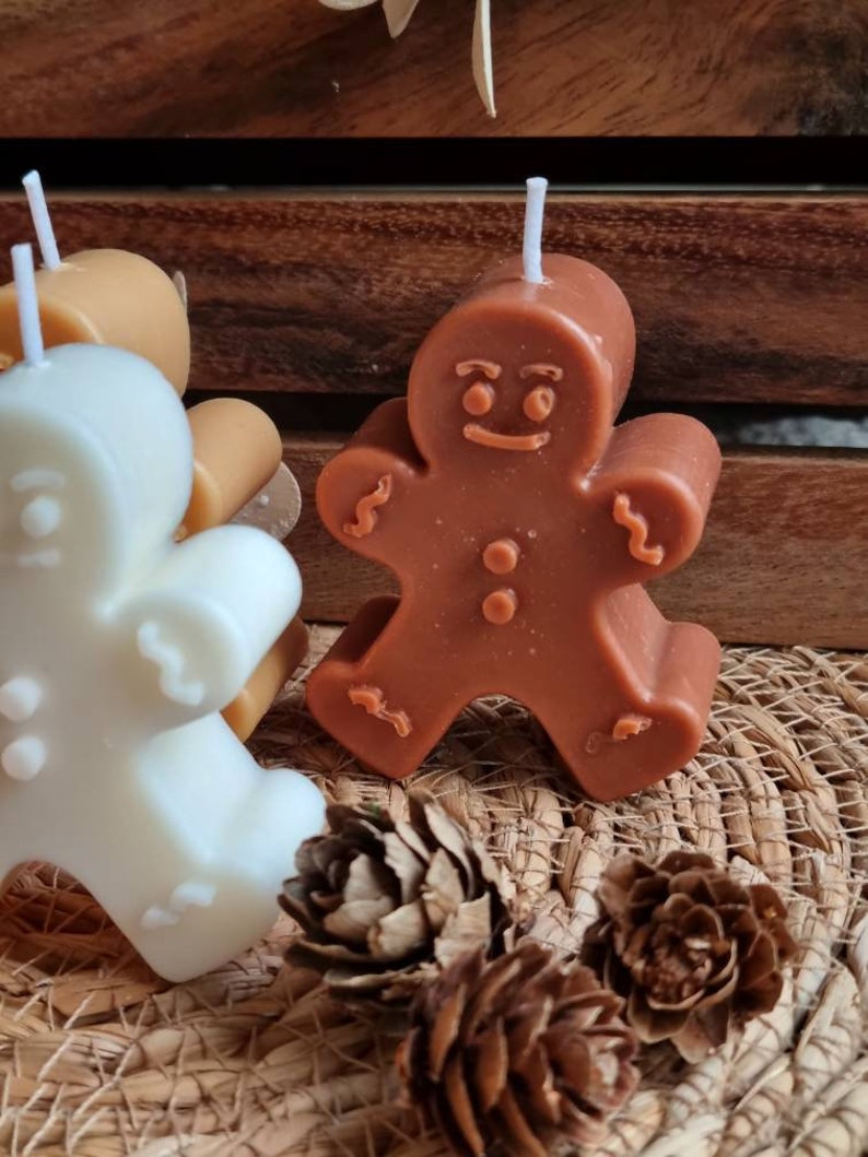 May include: Three gingerbread man shaped candles, one is brown, one is white, and one is tan. The candles are sitting on a woven basket surface with pine cones.