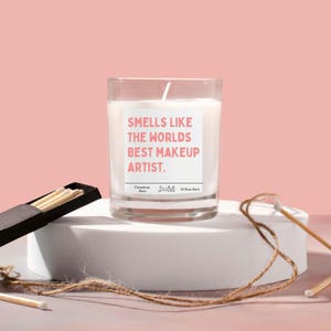 May include: A white candle with a pink label that reads "Smells like the world's best makeup artist." The candle is in a clear glass jar and is sitting on a white surface. There are matches in a black box and a piece of twine on the surface.