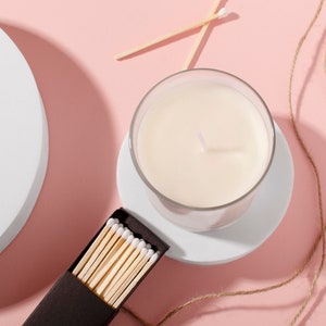 Puede incluir: Una vela blanca en un tarro de vidrio sobre un posavasos blanco, con una caja de cerillas y un trozo de cuerda sobre un fondo rosa.