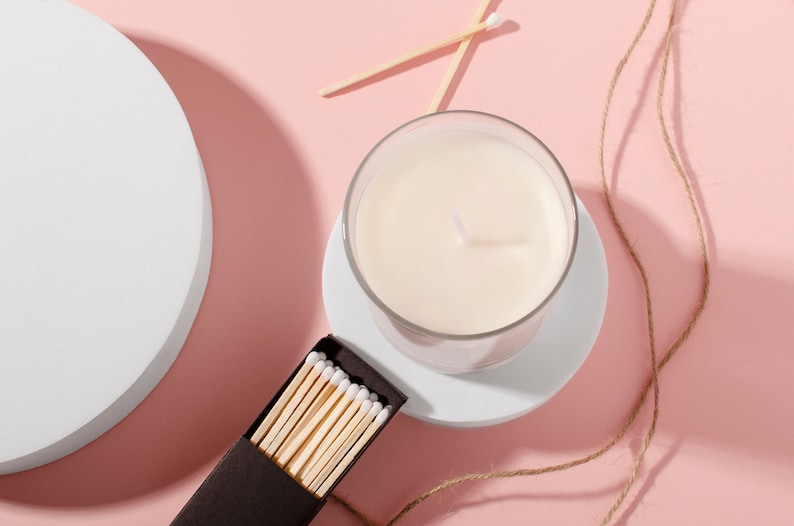 Puede incluir: Una vela blanca en un tarro de vidrio sobre un posavasos blanco, con una caja de cerillas y un trozo de cuerda sobre un fondo rosa.