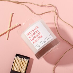 Puede incluir: Una vela blanca en un tarro de vidrio transparente con una etiqueta rosa que dice "Smells like you're my favourite solicitor." La vela est&aacute; sobre una superficie rosa con un cordel marr&oacute;n y cerillas.