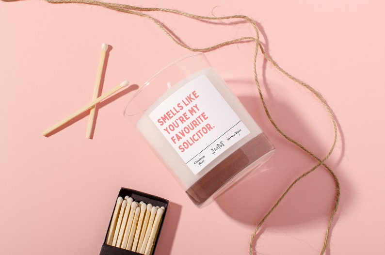 Puede incluir: Una vela blanca en un tarro de vidrio transparente con una etiqueta rosa que dice "Smells like you're my favourite solicitor." La vela est&aacute; sobre una superficie rosa con un cordel marr&oacute;n y cerillas.
