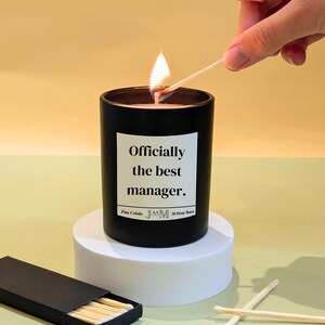May include: A black candle with the text "Officially the best manager" on a white label. The candle is lit with a match. The candle sits on a white pedestal. A box of matches is in the foreground.