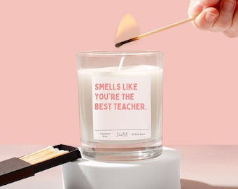 May include: A white candle with a pink label that reads "Smells like you're the best teacher." The candle is in a clear glass jar and is being lit with a match. The candle is on a white surface.