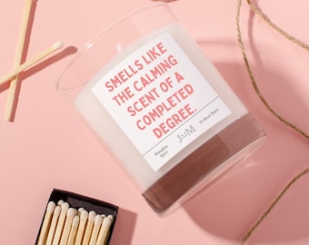 May include: A glass candle jar with a white label that reads "Smells like the calming scent of a completed degree." The candle is pumpkin spice scented and has a 30-hour burn time. A box of matches is in the lower left corner of the image.