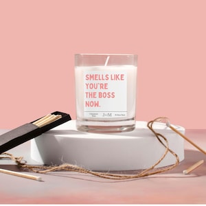 Może przedstawiać: Biała świeca w szklanym słoiku z różową etykietą, na której widnieje napis "Smells like you're the boss now." Świeca stoi na białym podłożu z pudełkiem zapałek i sznurkiem.