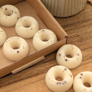 Könnte beinhalten: Eine Schachtel mit weißen, donut-förmigen Wachsschmelzen mit gesprenkelten Details. Die Schmelzen sind in einer braunen Pappschachtel auf einer Holzoberfläche angeordnet. Im Hintergrund ist ein Keramikbehälter zu sehen.