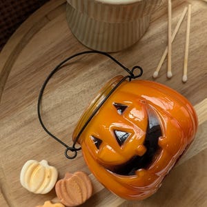 K&ouml;nnte beinhalten: Ein orangefarbener, k&uuml;rbisf&ouml;rmiger Kerzenhalter mit schwarzem Griff und einem geschnitzten Jack-o'-Lantern-Gesicht. Drei kleine, k&uuml;rbisf&ouml;rmige Seifen in Wei&szlig;-, Orange- und Braunt&ouml;nen befinden sich neben dem Kerzenhalter. Streichh&ouml;lzer und ein dekorativer St&auml;nder sind ebenfalls sichtbar.