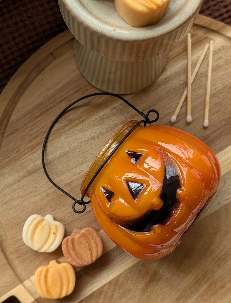 K&ouml;nnte beinhalten: Ein orangefarbener, k&uuml;rbisf&ouml;rmiger Kerzenhalter mit schwarzem Griff und einem geschnitzten Jack-o'-Lantern-Gesicht. Drei kleine, k&uuml;rbisf&ouml;rmige Seifen in Wei&szlig;-, Orange- und Braunt&ouml;nen befinden sich neben dem Kerzenhalter. Streichh&ouml;lzer und ein dekorativer St&auml;nder sind ebenfalls sichtbar.
