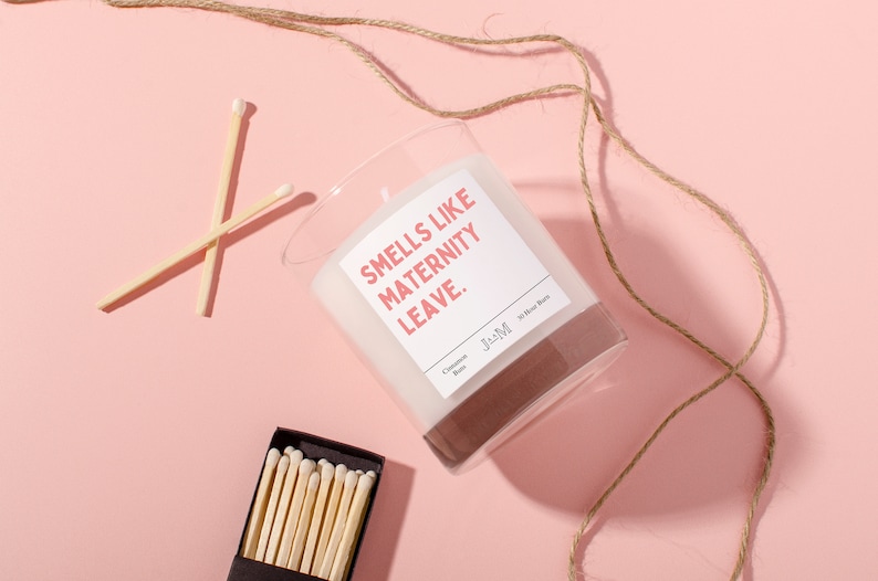 May include: A clear glass candle with the text "Smells like Maternity Leave" on a white label. The candle has a brown base and is accompanied by a box of matches and two loose matches. A length of twine is also present, all on a pink background.