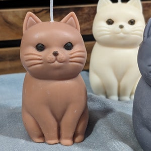 May include: Three cat-shaped candles, one brown, one white, and one gray. The brown cat candle has a white wick.
