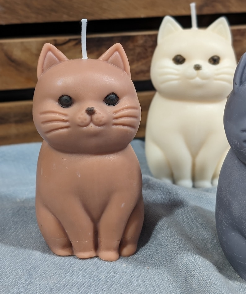 May include: Three cat-shaped candles, one brown, one white, and one gray. The brown cat candle has a white wick.